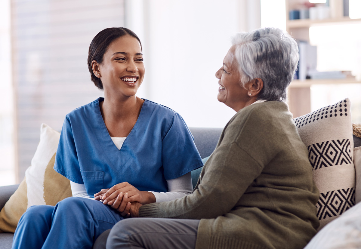 A professional caregiver in a warm-lit home setting gently supports an older adult who’s sitting in a comfortable chair; the caregiver is smiling, holding the person’s hand, creating a sense of trust and compassion.